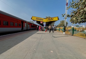 A train station platform with people walking towards a yellow overhead sign displaying the name Sehore in two languages. A red and grey passenger train is parked on the left side, and there are trees and a fence on the right side.