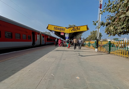 A train station platform with people walking towards a yellow overhead sign displaying the name Sehore in two languages. A red and grey passenger train is parked on the left side, and there are trees and a fence on the right side.