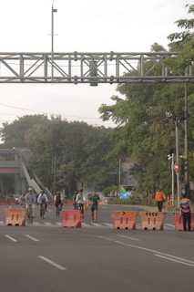 Several people are cycling and jogging along a street lined with orange barricades. The scene is set in a city area with greenery and trees in the background. Overhead, there is a metal structure likely used for signage or traffic lights. The atmosphere appears calm with a mix of commuters and joggers enjoying the open street.