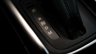 Close-up view of a car's automatic gear shift lever with visible letters indicating the gear positions: P, R, N, D, and M. The lever is made of black leather with stitching details and is surrounded by a metallic trim.
