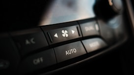 A close-up view of a car dashboard climate control panel, featuring buttons marked with A/C, fan speed, and auto mode. The panel is sleek and modern with a black finish and white text.