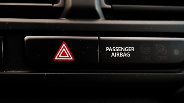 A close-up view of a car dashboard displaying an emergency hazard button with a red triangle symbol next to a passenger airbag indicator. The passenger airbag section has an icon indicating its on and off status.