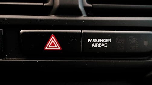A close-up view of a car dashboard displaying an emergency hazard button with a red triangle symbol next to a passenger airbag indicator. The passenger airbag section has an icon indicating its on and off status.