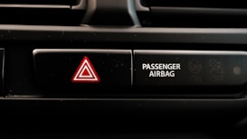 A close-up view of a car dashboard displaying an emergency hazard button with a red triangle symbol next to a passenger airbag indicator. The passenger airbag section has an icon indicating its on and off status.