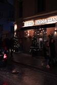 A dimly lit street scene features the exterior of a pizza restaurant named 'Pizza Napoletana'. The facade is adorned with festive lights and Christmas trees with decorations. People are walking past the pizzeria, and a car is partially visible on the wet road, suggesting recent rain or damp conditions.