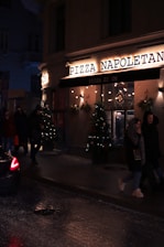 A dimly lit street scene features the exterior of a pizza restaurant named 'Pizza Napoletana'. The facade is adorned with festive lights and Christmas trees with decorations. People are walking past the pizzeria, and a car is partially visible on the wet road, suggesting recent rain or damp conditions.