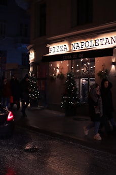 A dimly lit street scene features the exterior of a pizza restaurant named 'Pizza Napoletana'. The facade is adorned with festive lights and Christmas trees with decorations. People are walking past the pizzeria, and a car is partially visible on the wet road, suggesting recent rain or damp conditions.
