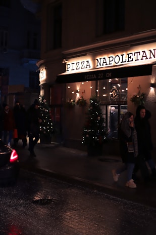 A dimly lit street scene features the exterior of a pizza restaurant named 'Pizza Napoletana'. The facade is adorned with festive lights and Christmas trees with decorations. People are walking past the pizzeria, and a car is partially visible on the wet road, suggesting recent rain or damp conditions.