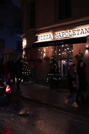 A dimly lit street scene features the exterior of a pizza restaurant named 'Pizza Napoletana'. The facade is adorned with festive lights and Christmas trees with decorations. People are walking past the pizzeria, and a car is partially visible on the wet road, suggesting recent rain or damp conditions.