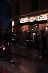 A dimly lit street scene features the exterior of a pizza restaurant named 'Pizza Napoletana'. The facade is adorned with festive lights and Christmas trees with decorations. People are walking past the pizzeria, and a car is partially visible on the wet road, suggesting recent rain or damp conditions.