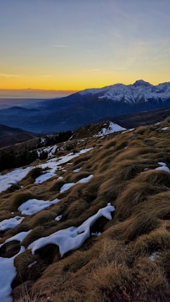 A breathtaking view of snow-capped mountains at sunset, with a foreground of rugged grassy hills partially covered in snow. The sky is a gradient of warm yellows blending into cool blues, creating a serene contrast with the mountains and valleys below.