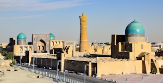 A historical cityscape featuring large domed structures with turquoise tiles and intricate Islamic architecture. In the background, a tall minaret rises prominently against the clear blue sky.