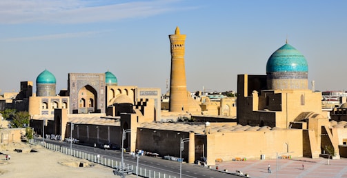 A historical cityscape featuring large domed structures with turquoise tiles and intricate Islamic architecture. In the background, a tall minaret rises prominently against the clear blue sky.
