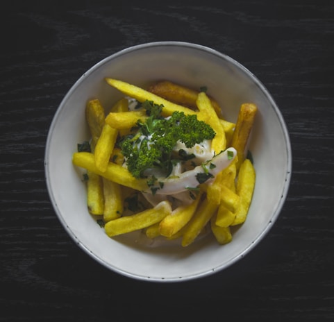 A bowl of golden French fries garnished with fresh green parsley, accompanied by a creamy white sauce, sitting on a dark wooden surface.