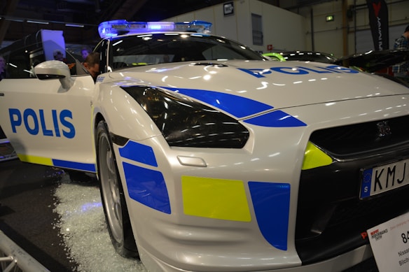 A white sports car marked as a police vehicle with 'Polis' written on it in blue letters. The car features yellow and blue geometric patterns and is equipped with a blue light bar on the roof. The image is taken indoors, possibly at an exhibition or show, as people are visible around the car with some standing with the car door open.