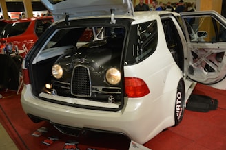 A white car with its trunk open, revealing what appears to be a vintage car front grille and headlights inside. The interior and doors are customized, with different color accents visible against a red carpet. Nearby, there are promotional materials and people in the background.
