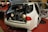 A white car with its trunk open, revealing what appears to be a vintage car front grille and headlights inside. The interior and doors are customized, with different color accents visible against a red carpet. Nearby, there are promotional materials and people in the background.