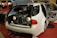 A white car with its trunk open, revealing what appears to be a vintage car front grille and headlights inside. The interior and doors are customized, with different color accents visible against a red carpet. Nearby, there are promotional materials and people in the background.