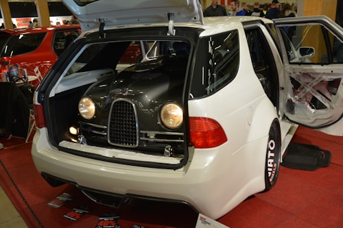 A white car with its trunk open, revealing what appears to be a vintage car front grille and headlights inside. The interior and doors are customized, with different color accents visible against a red carpet. Nearby, there are promotional materials and people in the background.