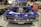 A classic blue car with a highly polished exterior and no hood, showcasing a powerful engine. The car is displayed indoors on a metallic checkered surface with spare tires and a red tool chest in the background. People are visible in the environment, suggesting a car exhibition or event.