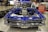 A classic blue car with a highly polished exterior and no hood, showcasing a powerful engine. The car is displayed indoors on a metallic checkered surface with spare tires and a red tool chest in the background. People are visible in the environment, suggesting a car exhibition or event.