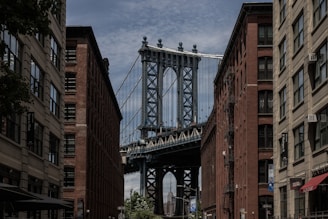 A view between two rows of brick buildings with the iconic steel structure of the Manhattan Bridge framed in the background. The sky is partly cloudy, and the urban setting includes street signs and architectural details typical of a city setting.