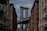 A view between two rows of brick buildings with the iconic steel structure of the Manhattan Bridge framed in the background. The sky is partly cloudy, and the urban setting includes street signs and architectural details typical of a city setting.