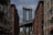 A view between two rows of brick buildings with the iconic steel structure of the Manhattan Bridge framed in the background. The sky is partly cloudy, and the urban setting includes street signs and architectural details typical of a city setting.