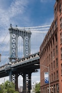 A large suspension bridge, featuring intricate steelwork, towers over an urban street scene. The left side shows the bridge's massive column and cables set against a clear blue sky. To the right, a red brick building stands with a banner advertising an event. Green foliage and streetlights are visible beneath the bridge, adding a touch of nature to the urban environment.