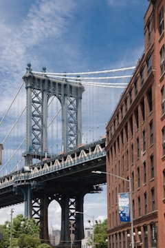 A large suspension bridge, featuring intricate steelwork, towers over an urban street scene. The left side shows the bridge's massive column and cables set against a clear blue sky. To the right, a red brick building stands with a banner advertising an event. Green foliage and streetlights are visible beneath the bridge, adding a touch of nature to the urban environment.