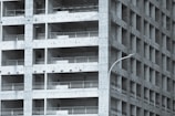 A partially constructed multi-story building with a grid-like structure. The concrete framework is visible, with repetitive square openings and vertical metal railings. A curved streetlamp is positioned near the building, adding an urban element to the scene.
