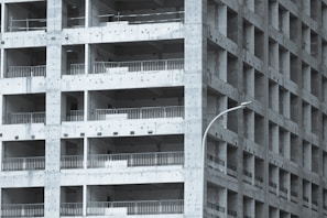 A partially constructed multi-story building with a grid-like structure. The concrete framework is visible, with repetitive square openings and vertical metal railings. A curved streetlamp is positioned near the building, adding an urban element to the scene.