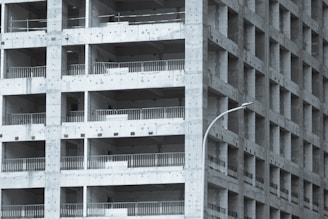 A partially constructed multi-story building with a grid-like structure. The concrete framework is visible, with repetitive square openings and vertical metal railings. A curved streetlamp is positioned near the building, adding an urban element to the scene.