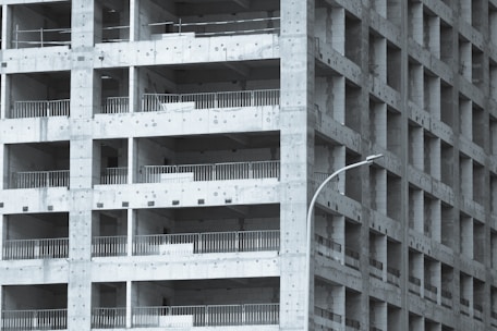 A partially constructed multi-story building with a grid-like structure. The concrete framework is visible, with repetitive square openings and vertical metal railings. A curved streetlamp is positioned near the building, adding an urban element to the scene.
