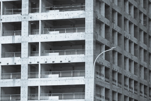 A partially constructed multi-story building with a grid-like structure. The concrete framework is visible, with repetitive square openings and vertical metal railings. A curved streetlamp is positioned near the building, adding an urban element to the scene.