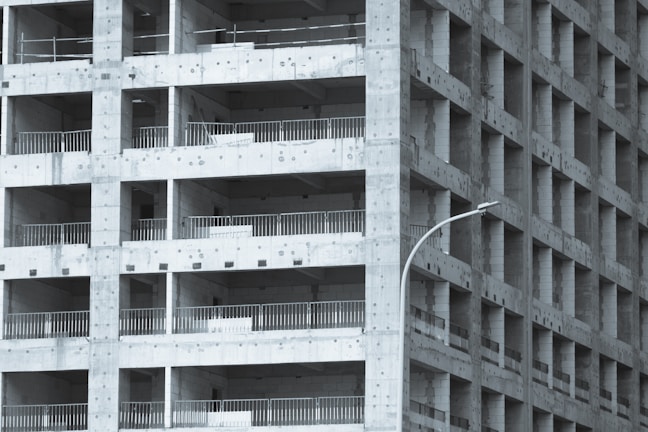 A partially constructed multi-story building with a grid-like structure. The concrete framework is visible, with repetitive square openings and vertical metal railings. A curved streetlamp is positioned near the building, adding an urban element to the scene.