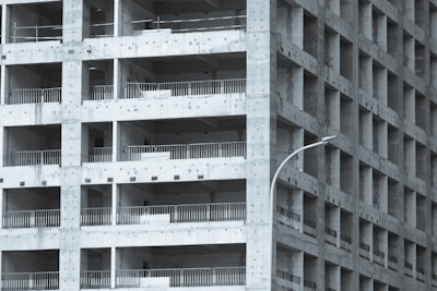 A partially constructed multi-story building with a grid-like structure. The concrete framework is visible, with repetitive square openings and vertical metal railings. A curved streetlamp is positioned near the building, adding an urban element to the scene.