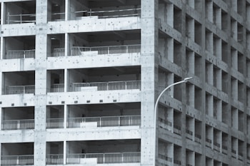 A partially constructed multi-story building with a grid-like structure. The concrete framework is visible, with repetitive square openings and vertical metal railings. A curved streetlamp is positioned near the building, adding an urban element to the scene.