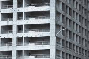 A partially constructed multi-story building with a grid-like structure. The concrete framework is visible, with repetitive square openings and vertical metal railings. A curved streetlamp is positioned near the building, adding an urban element to the scene.