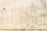 A plain beige wall with a small round vent near the top left corner. Two thin, horizontal wires run across the wall, casting shadows. A yellow horizontal pipe is attached at the bottom of the wall, with several small fittings visible along its length.