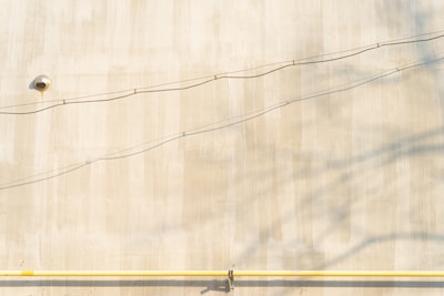 A plain beige wall with a small round vent near the top left corner. Two thin, horizontal wires run across the wall, casting shadows. A yellow horizontal pipe is attached at the bottom of the wall, with several small fittings visible along its length.