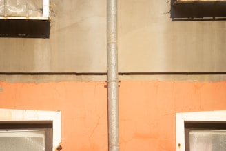 A vertical pipe runs down the center of a textured wall, which features two distinct color sections; the top half is gray and the bottom half is a faded orange. Below the pipe, there are two window frames with reflective surfaces, set into the wall.