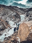 A group of people are trekking through a snowy mountain landscape. The path is rugged with rocks and ice, and the surrounding mountains are partially covered in snow. The sky is overcast, casting a muted light over the scene.