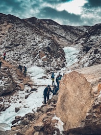 A group of people are trekking through a snowy mountain landscape. The path is rugged with rocks and ice, and the surrounding mountains are partially covered in snow. The sky is overcast, casting a muted light over the scene.