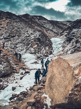 A group of people are trekking through a snowy mountain landscape. The path is rugged with rocks and ice, and the surrounding mountains are partially covered in snow. The sky is overcast, casting a muted light over the scene.