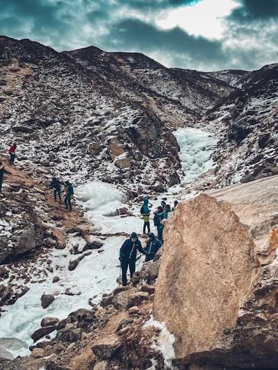 A group of people are trekking through a snowy mountain landscape. The path is rugged with rocks and ice, and the surrounding mountains are partially covered in snow. The sky is overcast, casting a muted light over the scene.