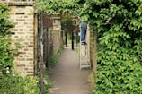An outdoor path is flanked by brick walls and heavy green foliage, leading to a house in the background. A person can be seen on a ladder trimming or managing the greenery on one side.