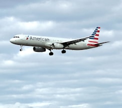 An airplane in flight with a cloudy sky background, displaying the logo and colors of an airline on its fuselage and tail. The landing gear is visible, indicating it is either taking off or preparing to land.