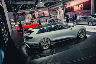 A slick, modern exhibition space featuring several high-end cars. A white sedan is prominently displayed on a circular platform, surrounded by other luxury vehicles in various colors, including a striking red car. The environment is bustling with people, likely attendees and exhibitors, moving around the cars. Large digital screens and branded banners adorn the background.