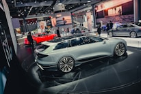 A slick, modern exhibition space featuring several high-end cars. A white sedan is prominently displayed on a circular platform, surrounded by other luxury vehicles in various colors, including a striking red car. The environment is bustling with people, likely attendees and exhibitors, moving around the cars. Large digital screens and branded banners adorn the background.